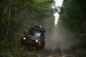 A rugged off-road vehicle is navigating a narrow, muddy path surrounded by dense green forest. The vehicle is loaded with gear on its roof, indicating an adventurous journey. Mist or dust creates a hazy atmosphere, adding to the sense of wilderness.