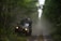 A rugged off-road vehicle is navigating a narrow, muddy path surrounded by dense green forest. The vehicle is loaded with gear on its roof, indicating an adventurous journey. Mist or dust creates a hazy atmosphere, adding to the sense of wilderness.
