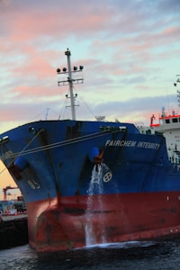 A large cargo ship, named the Fairchem Integrity, is docked at a port. Water is seen flowing from the side of the vessel near the front. The sky is filled with pink and blue hues typical of either a sunrise or sunset. The ship's hull is dark blue with some red near the waterline.