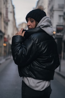 A person stands in a narrow urban street wearing a black beanie with a fur pom-pom, black puffer jacket, and gray hoodie. The street is lined with buildings, and there is soft lighting, suggesting a cloudy day.
