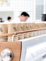 Paper coffee cups with a minimalist design featuring a percentage symbol on them are neatly arranged in rows atop a modern espresso machine. The background shows blurred figures, likely baristas, in a clean, bright cafe setting.
