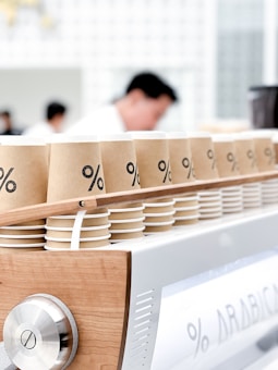 Paper coffee cups with a minimalist design featuring a percentage symbol on them are neatly arranged in rows atop a modern espresso machine. The background shows blurred figures, likely baristas, in a clean, bright cafe setting.