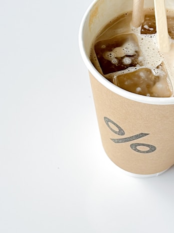 A brown paper cup with a percentage symbol printed on it, filled with iced coffee. Ice cubes are visible on the surface, and a wooden stirrer is placed inside the drink. The cup is set on a light background, offering a minimalist and clean appearance.