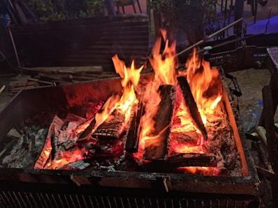 A vibrant fire burns in a rectangular metal container, with large flames licking upward and glowing embers scattered among charred logs. The surrounding area is dimly lit, and there are hints of outdoor furniture and foliage in the background.