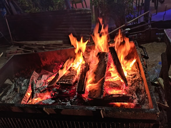 A vibrant fire burns in a rectangular metal container, with large flames licking upward and glowing embers scattered among charred logs. The surrounding area is dimly lit, and there are hints of outdoor furniture and foliage in the background.