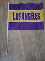 A small flag with the words 'Los Angeles' written in bold purple letters on a yellow background, surrounded by a purple design. The flag is attached to a wooden stick and is placed on a wooden floor.