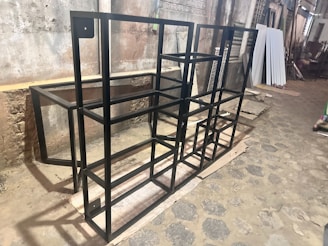 The image depicts a metal framework structure set on a rough, cobblestone floor in what appears to be a workshop or industrial setting. The walls are aged and stained, and various building materials and tools can be seen in the background. The metal framework is black and consists of multiple rectangular shapes and segments.