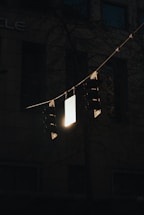 A set of dark traffic lights is silhouetted against a dimly lit urban background. The lights hang from horizontal wires with a bright, reflective sign in the center catching some light. The surrounding area is shadowy, with subtle hints of a building in the backdrop.