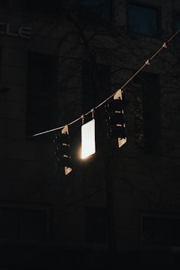 A set of dark traffic lights is silhouetted against a dimly lit urban background. The lights hang from horizontal wires with a bright, reflective sign in the center catching some light. The surrounding area is shadowy, with subtle hints of a building in the backdrop.
