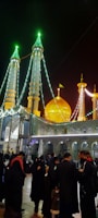 A nighttime scene features a beautifully illuminated mosque with multiple minarets adorned with green and white lights. A golden dome stands prominently amidst the structures, which are intricately decorated with floral and geometric patterns. People gather in the courtyard, dressed in traditional attire, creating a sense of community.
