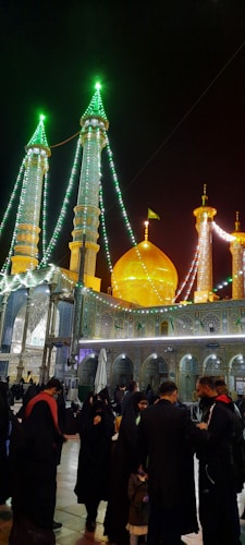 A nighttime scene features a beautifully illuminated mosque with multiple minarets adorned with green and white lights. A golden dome stands prominently amidst the structures, which are intricately decorated with floral and geometric patterns. People gather in the courtyard, dressed in traditional attire, creating a sense of community.