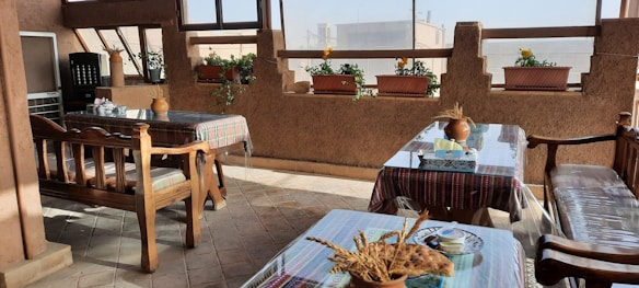 A cozy indoor seating area with wooden benches and chairs, covered in tablecloths with plaid patterns. The tables have decorative items such as vases with dried plants. Along the walls, there are large windows and several potted plants with flowers placed on a shelf, allowing natural light to illuminate the space.