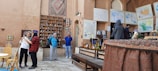 A group of people are in a cultural or historical indoor setting with earthy-toned walls adorned with maps and fabric hangings. Shelves display pottery and souvenirs. A table and chairs are in the foreground. Several individuals are interacting, taking photos, and observing the surroundings.