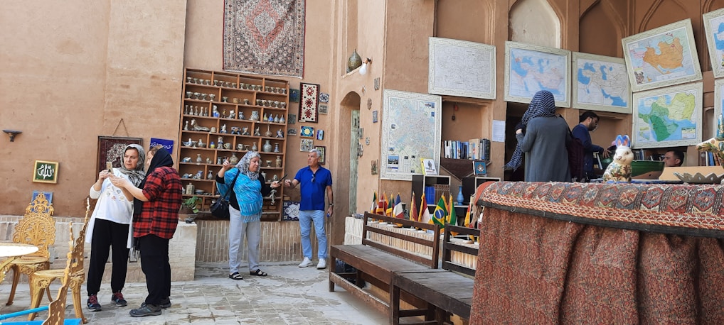 A group of people are in a cultural or historical indoor setting with earthy-toned walls adorned with maps and fabric hangings. Shelves display pottery and souvenirs. A table and chairs are in the foreground. Several individuals are interacting, taking photos, and observing the surroundings.