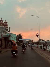 A street scene at sunset where several people are riding motorbikes along a road. The sun is low in the sky, casting an orange glow. Buildings and street lights line the road, and the sky transitions from blue to orange as the sun sets.
