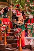 A festive scene featuring cartoon characters dressed in holiday attire with red and green outfits, standing next to a decorated Christmas tree with lights and ornaments. The setting includes garlands and large wrapped gifts in the background.