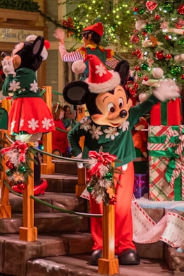 A festive scene featuring cartoon characters dressed in holiday attire with red and green outfits, standing next to a decorated Christmas tree with lights and ornaments. The setting includes garlands and large wrapped gifts in the background.