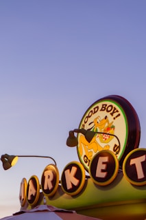 A colorful retro-style market sign displays the words 'Good Boy!' with a cartoon dog carrying a basket of groceries. The sign is illuminated, with individual letters of 'MARKET' circled and lit from behind. Two lamp fixtures are visible, adding to the vintage aesthetic against a clear evening sky.