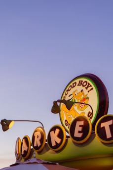 A colorful retro-style market sign displays the words 'Good Boy!' with a cartoon dog carrying a basket of groceries. The sign is illuminated, with individual letters of 'MARKET' circled and lit from behind. Two lamp fixtures are visible, adding to the vintage aesthetic against a clear evening sky.
