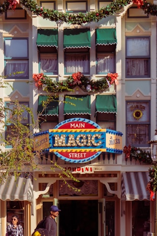 An ornate facade of a building decorated with garlands and red bows, featuring a vintage-style sign that reads 'Main Magic Street' in bright colors. The windows are adorned with green awnings, and there are people walking by on the street below.