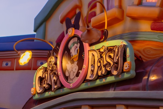 A vibrant sign featuring a cartoon duck character dressed as a chef, holding a whisk, with the words 'Caf&eacute; Daisy' elegantly designed. The background displays colorful architecture with whimsical design elements.