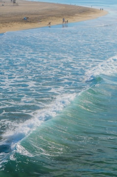 A serene beach scene with gentle waves crashing on the shore. In the foreground, the ocean water is a vivid turquoise, transitioning to a darker blue towards the horizon. The sandy beach extends along the upper third of the image, where a few people are seen walking and enjoying the sunny day. The blue sky complements the sea, creating a calming atmosphere.
