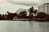 A scenic urban lakeside view with prominent hotels in the background. The foreground features calm water, while lush greenery and tall palm trees line the shoreline. A small red-roofed gazebo sits on a pier, adding charm to the landscape. The sky is overcast, giving the scene a moody ambiance.