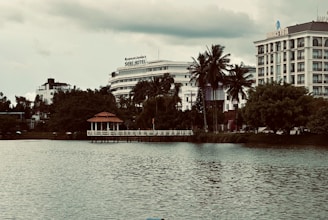 A scenic urban lakeside view with prominent hotels in the background. The foreground features calm water, while lush greenery and tall palm trees line the shoreline. A small red-roofed gazebo sits on a pier, adding charm to the landscape. The sky is overcast, giving the scene a moody ambiance.