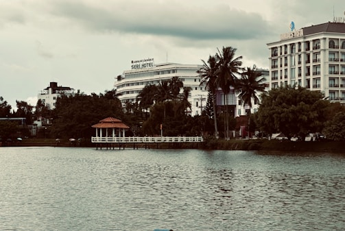A scenic urban lakeside view with prominent hotels in the background. The foreground features calm water, while lush greenery and tall palm trees line the shoreline. A small red-roofed gazebo sits on a pier, adding charm to the landscape. The sky is overcast, giving the scene a moody ambiance.