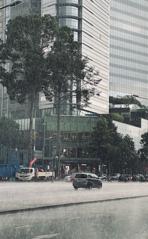 Heavy rain falls on a busy urban street with a large modern glass building in the background. Cars and motorcycles are moving through the wet street while tree leaves are visibly damp. The overall scene captures the hustle of city life during a rainstorm.
