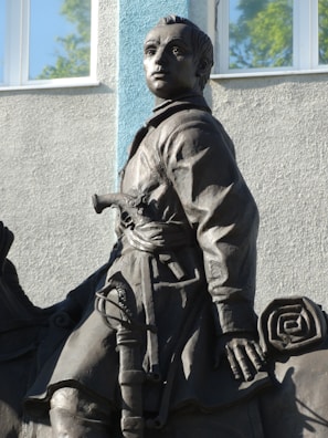A bronze statue of a historical figure riding a horse, holding a coiled scroll in one hand and wearing traditional attire with a sword belt. The figure gazes ahead with a determined expression. The background includes a building with a large window, featuring a light blue and gray color scheme.