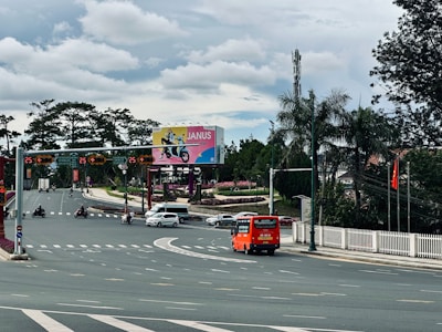 A busy intersection with several vehicles including an orange bus and motorbikes on the road. There is a large billboard displaying an advertisement in the background, surrounded by greenery and palm trees. Traffic lights and street signs are visible, and a Vietnamese flag is flying on the right.