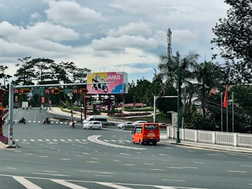 A busy intersection with several vehicles including an orange bus and motorbikes on the road. There is a large billboard displaying an advertisement in the background, surrounded by greenery and palm trees. Traffic lights and street signs are visible, and a Vietnamese flag is flying on the right.