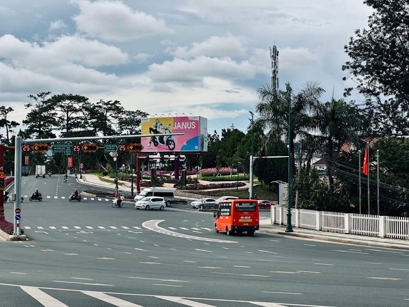 A busy intersection with several vehicles including an orange bus and motorbikes on the road. There is a large billboard displaying an advertisement in the background, surrounded by greenery and palm trees. Traffic lights and street signs are visible, and a Vietnamese flag is flying on the right.