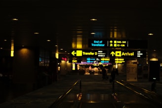 Dimly lit airport corridor with illuminated signs indicating directions for transfer, arrivals, and gates. People can be seen walking in the background.