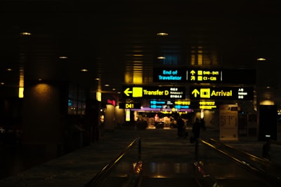 Dimly lit airport corridor with illuminated signs indicating directions for transfer, arrivals, and gates. People can be seen walking in the background.