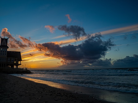 A dramatic sunset over the ocean with a silhouette of a large house on the left. Dark clouds float across the colorful sky with shades of orange and pink, while the waves gently crash onto the sandy beach.