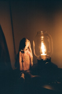 A plush rabbit toy wearing a coat is seated in a dimly lit room next to a glass lamp emitting a warm glow.