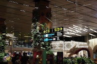 An indoor area bustling with activity, featuring signage directing towards different terminal gates, shopping and lounges. The space is adorned with lush greenery and illuminated by modern lighting fixtures. Visible storefronts include luxury brands like Dior and Hennessy. The setting includes decorative elements like plant-covered pillars and a large sculptural piece.
