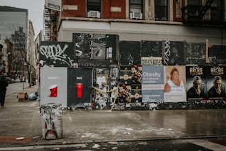 A city street corner featuring a graffiti-covered wall with various posters and advertisements. The ads include 'naughty or nice' themed cups, obesity awareness, and promotional material for a show called 'Archie.' The area appears slightly cluttered with a trash can, boxes, and scattered debris on a wet sidewalk.