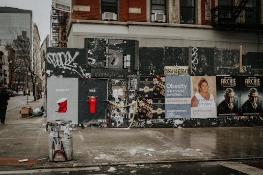 A city street corner featuring a graffiti-covered wall with various posters and advertisements. The ads include 'naughty or nice' themed cups, obesity awareness, and promotional material for a show called 'Archie.' The area appears slightly cluttered with a trash can, boxes, and scattered debris on a wet sidewalk.