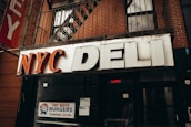 A storefront sign reading 'NYC Deli' in large, bold letters. Beneath the sign, a poster advertises 'Fat Boys Burgers Coming Soon', featuring an illustration of a cartoon character. The storefront is set against a brick facade with a metal balcony railing and fire escape above.