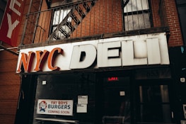 A storefront sign reading 'NYC Deli' in large, bold letters. Beneath the sign, a poster advertises 'Fat Boys Burgers Coming Soon', featuring an illustration of a cartoon character. The storefront is set against a brick facade with a metal balcony railing and fire escape above.