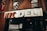 A storefront sign reading 'NYC Deli' in large, bold letters. Beneath the sign, a poster advertises 'Fat Boys Burgers Coming Soon', featuring an illustration of a cartoon character. The storefront is set against a brick facade with a metal balcony railing and fire escape above.