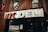 A storefront sign reading 'NYC Deli' in large, bold letters. Beneath the sign, a poster advertises 'Fat Boys Burgers Coming Soon', featuring an illustration of a cartoon character. The storefront is set against a brick facade with a metal balcony railing and fire escape above.