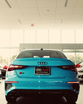 A bright blue Audi S3 is parked inside a car dealership showroom. The vehicle's rear end is prominently displayed, highlighting its sleek aerodynamic design and modern taillights. The dealership is well-lit with large windows that let in natural light, and several other cars are visible in the background.