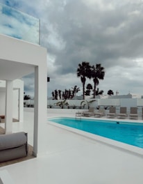 A modern outdoor pool area with a clear blue swimming pool surrounded by white architectural structures. There are several beige lounge chairs neatly lined up beside the pool. In the background, tall palm trees and a cloudy sky create a dramatic contrast with the bright surroundings.