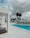 A modern outdoor pool area with a clear blue swimming pool surrounded by white architectural structures. There are several beige lounge chairs neatly lined up beside the pool. In the background, tall palm trees and a cloudy sky create a dramatic contrast with the bright surroundings.