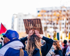 A crowd of people is gathered at an outdoor event, with one person holding a cardboard sign that reads 'HUMANITY NOW'. The scene includes various individuals wearing casual clothing, some with hats and masks. The background features urban elements and blurred foliage, suggesting a public demonstration or protest.