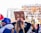 A crowd of people is gathered at an outdoor event, with one person holding a cardboard sign that reads 'HUMANITY NOW'. The scene includes various individuals wearing casual clothing, some with hats and masks. The background features urban elements and blurred foliage, suggesting a public demonstration or protest.
